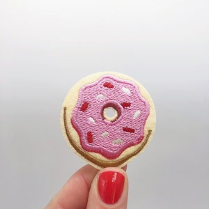 May include: An embroidered donut patch. The patch features a pink iced donut with red and white sprinkles. The base is beige with a brown outline and a smiling face.
