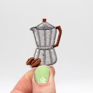 Può includere: Un toppa ricamata a forma di caffettiera di ferro, di colore argento e marrone. La toppa è su uno sfondo bianco.