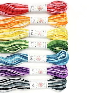 May include: Seven spools of embroidery floss in a rainbow of colours, including red, orange, yellow, green, blue, purple, and grey. Each spool has a white label with a flower design and a number printed on it: 634, 628, 644, 679, 699, 612, and 654.