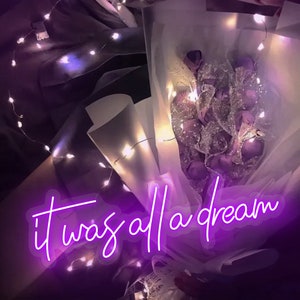 May include: A bouquet of purple flowers wrapped in white tissue paper with a string of white fairy lights wrapped around it. The text "it was all a dream" is written in neon purple letters.