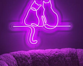 May include: A bright purple neon sign featuring a continuous line drawing of two cats sitting side by side on a horizontal line. The cats are depicted in profile, with one cat's long tail hanging down below the line, creating a glowing wall light.