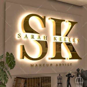 May include: A wall-mounted sign featuring large, gold-colored, three-dimensional letters S and K with a warm, backlit glow. Below the letters, the text SARAH KELLEY is displayed in gold, followed by the text MAKEUP ARYIST in smaller gold lettering.