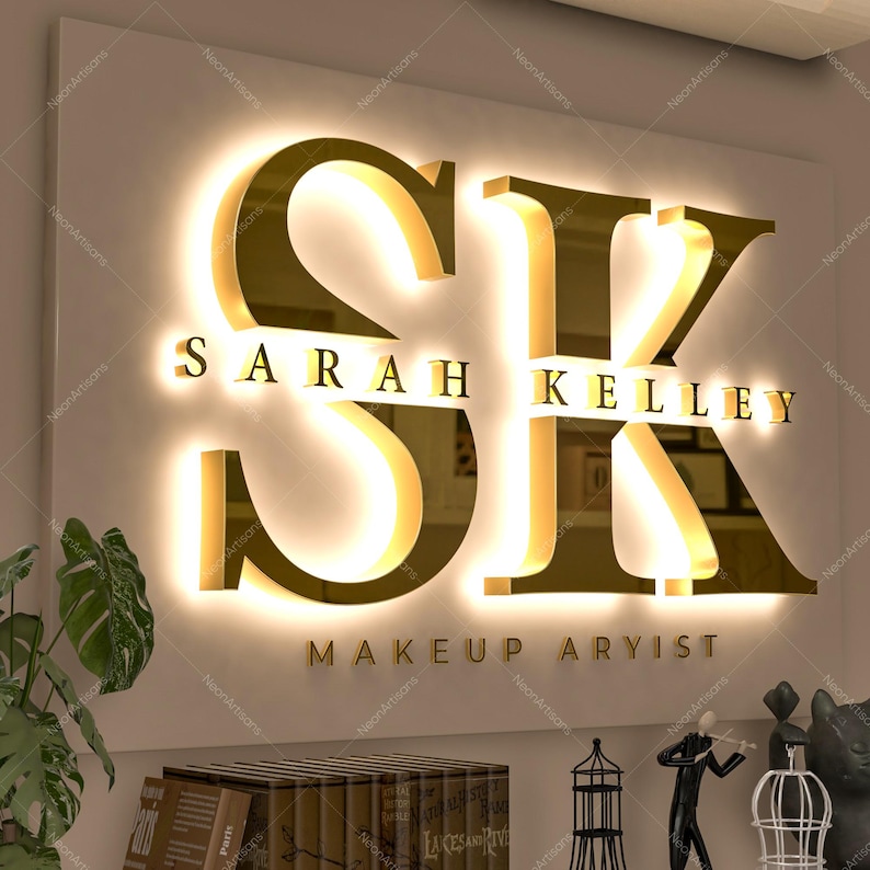 May include: A wall-mounted sign featuring large, gold-colored, three-dimensional letters S and K with a warm, backlit glow. Below the letters, the text SARAH KELLEY is displayed in gold, followed by the text MAKEUP ARYIST in smaller gold lettering.