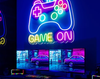Può includere: Un cartello al neon con un controller per videogiochi colorato e le parole "GAME ON" in giallo e rosa. Il cartello è montato su una parete sopra due monitor per computer con uno sfondo scuro.