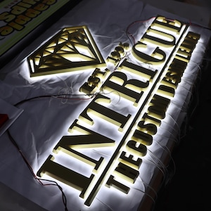 May include: Gold-colored metal sign with the word "INTrigue" in large letters and the text "The Custom Letter" in smaller letters below. The sign is illuminated with white LED lights and features a diamond shape above the word "INTrigue".