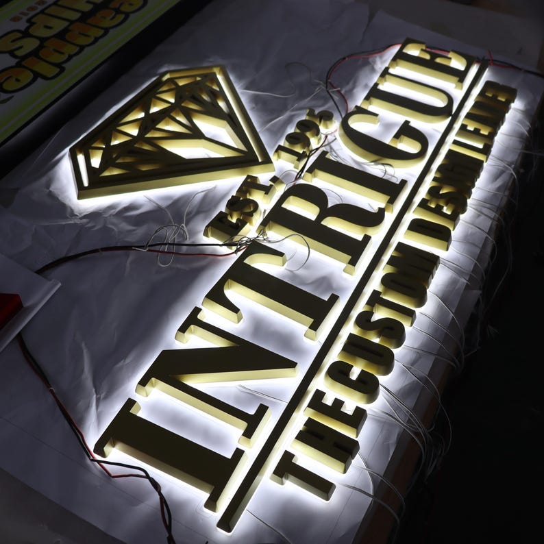 May include: Gold-colored metal sign with the word "INTrigue" in large letters and the text "The Custom Letter" in smaller letters below. The sign is illuminated with white LED lights and features a diamond shape above the word "INTrigue".