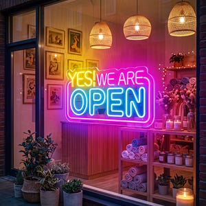 Könnte beinhalten: Ein Schaufenster mit einem hellen Neonschild, das YES! WE ARE OPEN in Gelb, Pink und Blau anzeigt. Im Inneren befinden sich Holzregale mit gerollten Handtüchern, Kerzen und Trockenblumen, die von drei geflochtenen Pendelleuchten beleuchtet werden.