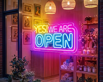 May include: A storefront window display featuring a bright neon sign that reads YES! WE ARE OPEN in yellow, pink, and blue. The interior shows wooden shelving units stocked with rolled towels, candles, and dried flowers, illuminated by three woven pendant lights.