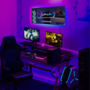 以下が含まれることがあります： 黒と青のゲーミングチェア、黒いコンピュータタワー、2つのモニター、キーボードが置かれたデスクがあるゲーミングセットアップ。デスクの後ろの壁には「Olivia's Bedroom」と書かれたネオンサインがある。