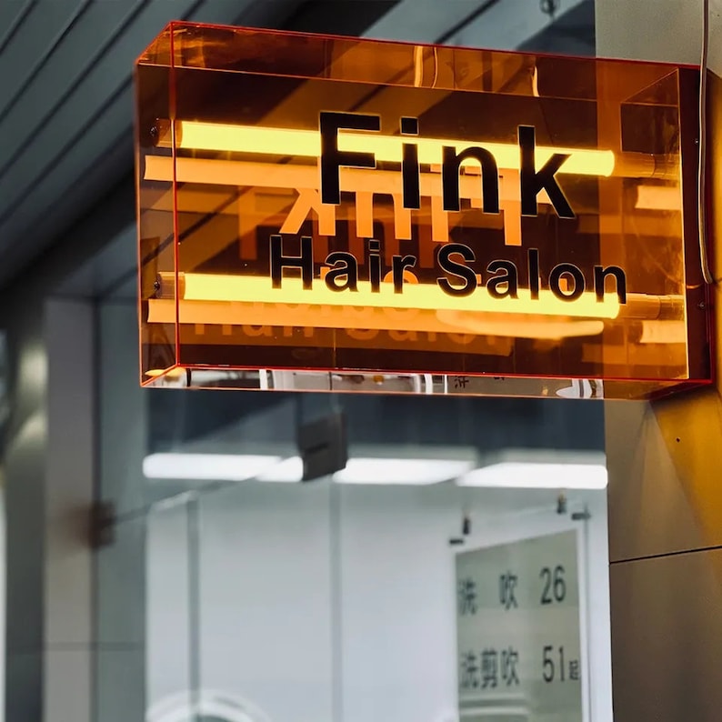 May include: A rectangular orange sign with the text "Fink Hair Salon" in black lettering. The sign is illuminated with yellow fluorescent lights.