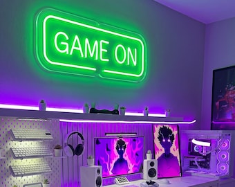 Könnte beinhalten: Ein rechteckiges Neonschild mit hellgrünem Licht und der Aufschrift GAME ON, das an einer Wand über einem Schreibtisch montiert ist. Der Arbeitsplatz umfasst einen weißen Computer-Tower, zwei Monitore mit violetten Anime-Grafiken und weiße Lautsprecher.