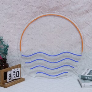 Puede incluir: Un letrero de ne&oacute;n que representa un sol naranja estilizado poni&eacute;ndose sobre olas azules. El letrero est&aacute; montado en una pared blanca.