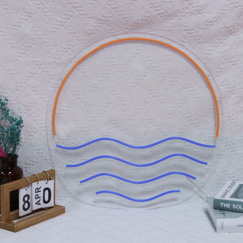 Puede incluir: Un letrero de ne&oacute;n que representa un sol naranja estilizado poni&eacute;ndose sobre olas azules. El letrero est&aacute; montado en una pared blanca.