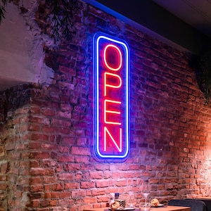 Könnte beinhalten: Ein vertikales, rechteckiges Neonschild an einer rustikalen Ziegelwand. Das Schild zeigt das Wort OPEN in leuchtend roten Neonbuchstaben, umschlossen von einem durchgehenden blauen Neonrahmen, der der abgerundeten rechteckigen Form folgt.