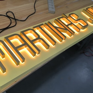 May include: A rectangular sign with a light yellow base featuring raised, metallic gold-toned letters that spell out MARTINA'S JUS. The letters are backlit with a warm, glowing light, creating a distinct silhouette against the yellow panel.