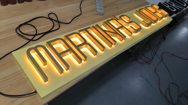 May include: A rectangular sign with a light yellow base featuring raised, metallic gold-toned letters that spell out MARTINA'S JUS. The letters are backlit with a warm, glowing light, creating a distinct silhouette against the yellow panel.