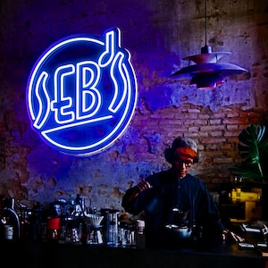 May include: A blue neon sign with the text "SEB's" and a musical note in a circle, hanging on a brick wall. A person wearing a black shirt and a red hat is making coffee behind a counter.
