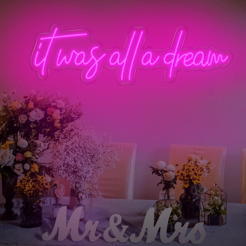 May include: Pink neon sign with the text "it was all a dream" hanging on a wall above a table with white flowers and a sign that says "Mr & Mrs".