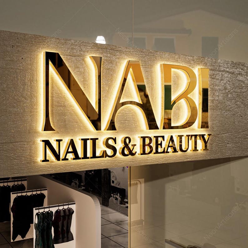 May include: Gold-colored sign with the word "NABI" in large letters and the words "NAILS & BEAUTY" in smaller letters below. The sign is illuminated with white lights.