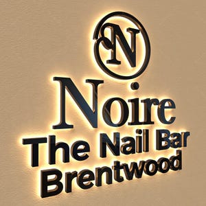 May include: Black illuminated sign for "Noire The Nail Bar Brentwood". The sign features a circular logo with the letter "N" and the business name in a bold, sans-serif font. The sign is mounted on a beige wall.