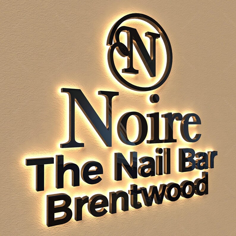 May include: Black illuminated sign for "Noire The Nail Bar Brentwood". The sign features a circular logo with the letter "N" and the business name in a bold, sans-serif font. The sign is mounted on a beige wall.
