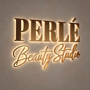 May include: A three-dimensional wall sign featuring the word PERL&Eacute; in large, gold-toned serif capital letters, positioned above the words Beauty Studio in a gold-toned cursive script, both illuminated from behind with a warm glow.