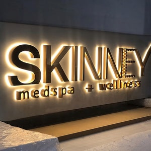 May include: A three-dimensional wall sign featuring the word SKINNEY in large, dark, backlit capital letters, with the words medspa + wellness in smaller, matching backlit lowercase letters positioned directly below the main text.
