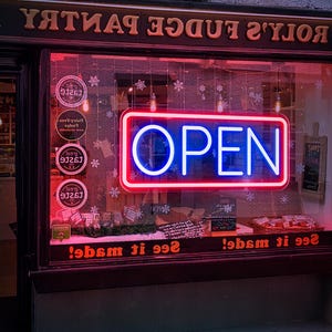 May include: A red and blue neon sign with the word "OPEN" in white letters. The sign is in a storefront window with the words "See it made!" written below it.