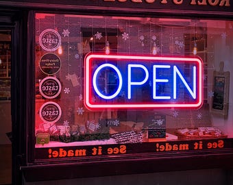 May include: A red and blue neon sign with the word "OPEN" in white letters. The sign is in a storefront window with the words "See it made!" written below it.