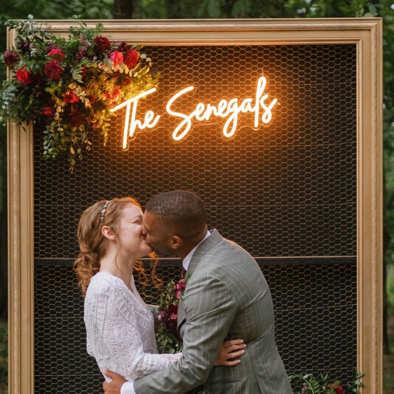 Bride and groom sharing a kiss in front of a custom wedding neon sign that reads 'The Senegals' with an elegant frame surrounded by lush red and burgundy florals, Perfect for couples seeking inspiration for their own unique wedding signage