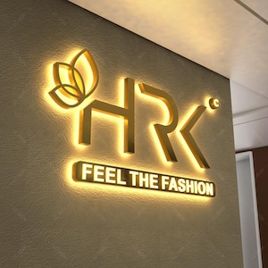 May include: A three-dimensional wall sign featuring the gold-colored letters HRK and a stylized leaf icon. Below the letters, a rectangular gold sign displays the text FEEL THE FASHION. The entire display is illuminated with a warm glow.
