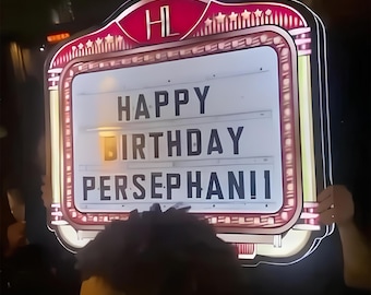 May include: A vintage-style illuminated sign with a red and gold frame displays the message "HAPPY BIRTHDAY PERSEPHANII" in bold, white letters. The sign has a retro aesthetic, perfect for a birthday celebration.