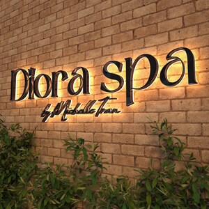 May include: Illuminated "Diora Spa" sign with the text "by Michelle Tran" on a brick wall. The sign features black lettering with a warm, glowing backlight. Green plants are visible at the bottom of the image.