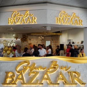 May include: A gold-colored sign with the text "Glam Bar Bazaar" in a cursive font. The sign is illuminated with lights and is mounted on a white brick wall.