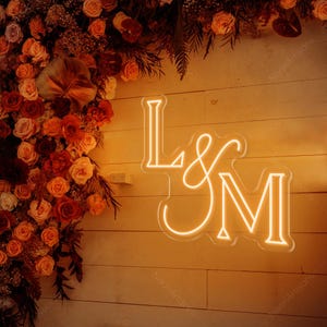 May include: A neon sign displaying the letters "L & M" in a warm, glowing orange hue. The sign is set against a backdrop of a wooden wall and a floral arrangement of roses and other flowers in shades of red, orange, and yellow.