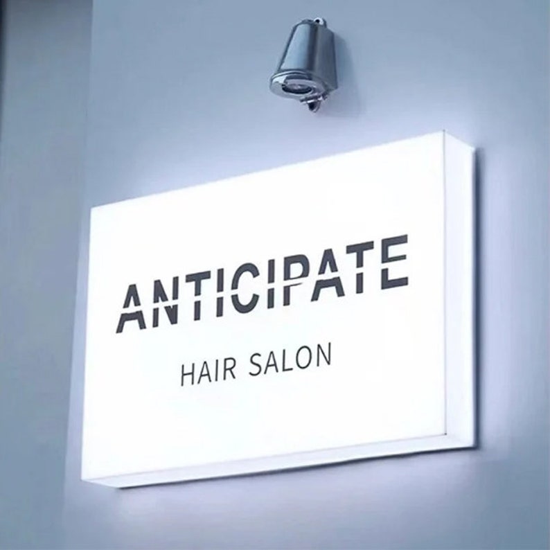 May include: A white rectangular sign with black text that reads "ANTICIPATE HAIR SALON" mounted on a white wall. A silver light fixture is mounted above the sign.