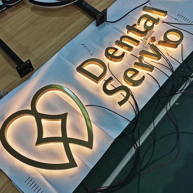 May include: A lit-up sign for "Dental Servio" in gold lettering, with a heart-shaped logo. The sign is on a white surface with wiring visible. The letters are illuminated with a warm glow.