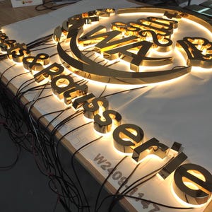 May include: A golden, illuminated sign for a restaurant, featuring the words "Restaurant Rotisserie" in a stylized font. The sign is composed of individual, three-dimensional letters with internal lighting, and is likely for outdoor use.