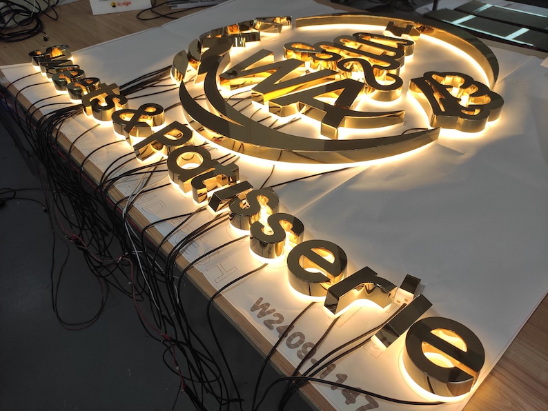 May include: A golden, illuminated sign for a restaurant, featuring the words "Restaurant Rotisserie" in a stylized font. The sign is composed of individual, three-dimensional letters with internal lighting, and is likely for outdoor use.
