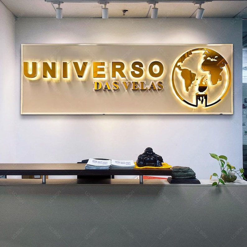 May include: A gold sign with the text "UNIVERSO DAS VELAS" and a gold globe graphic. The sign is mounted on a white wall above a dark brown counter.