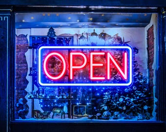Könnte beinhalten: Ein rotes Neon-Schild mit blauem Rand, auf dem in einem Schaufenster "GEÖFFNET" steht. Das Schaufenster ist mit Weihnachtslichtern und einem kleinen Weihnachtsbaum geschmückt.