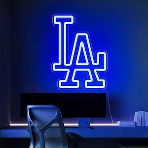 Pode incluir: Uma placa de néon azul com as letras "LA" em contorno branco. A placa está montada em uma parede acima de uma mesa com um monitor de computador e uma cadeira.