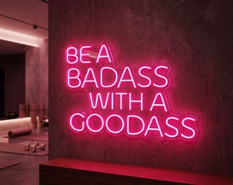 May include: A neon sign in bright pink, reading "BE A BADASS WITH A GOODASS", against a textured grey wall. The sign is above a dark wood bench. The room has a modern, minimalist design with a yoga mat visible.
