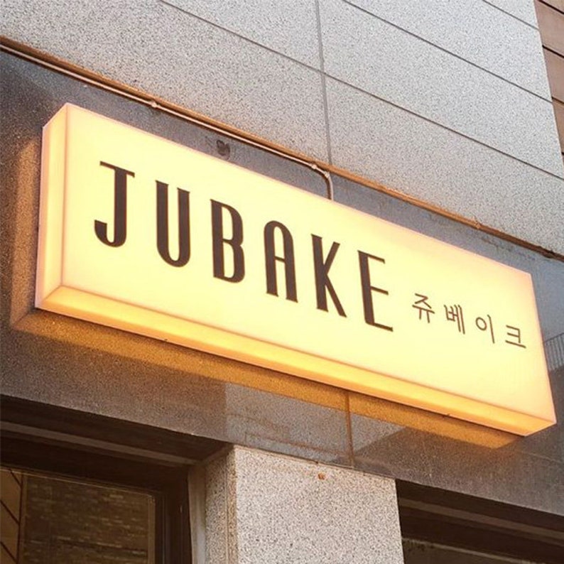 May include: A white illuminated sign with black lettering that reads "JUBAKe" and "쥬베이크" in Korean. The sign is mounted on a building.