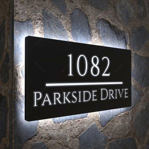 Pode incluir: Uma placa de endereço retangular preta com o número "1082" e as palavras "PARKSIDE DRIVE" em letras brancas iluminadas. A placa está montada em uma parede de pedra e tem uma borda branca brilhante.