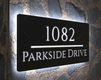 Puede incluir: Un letrero de dirección rectangular negro con el número "1082" y las palabras "PARKSIDE DRIVE" en letras blancas iluminadas. El letrero está montado en una pared de piedra y tiene un borde blanco brillante.
