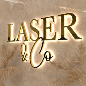 May include: A three-dimensional wall sign featuring the gold-colored text LASER & Co. The letters have a polished metallic finish and are illuminated from behind with a warm white glow, creating a halo effect against the wall surface.