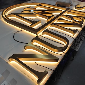 May include: Illuminated gold-colored metal signage spelling out "UNIS". The letters are three-dimensional and have a warm, glowing light emanating from within. The sign is resting on a white surface, with wires visible.