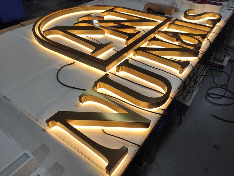 May include: Illuminated gold-colored metal signage spelling out "UNIS". The letters are three-dimensional and have a warm, glowing light emanating from within. The sign is resting on a white surface, with wires visible.