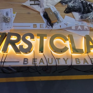 May include: A gold-toned metal sign featuring the words FIRST CLASS in large, illuminated 3D letters with a warm glow, positioned above the text BEAUTY BAR in smaller, non-illuminated letters, with black electrical wires extending from the base.